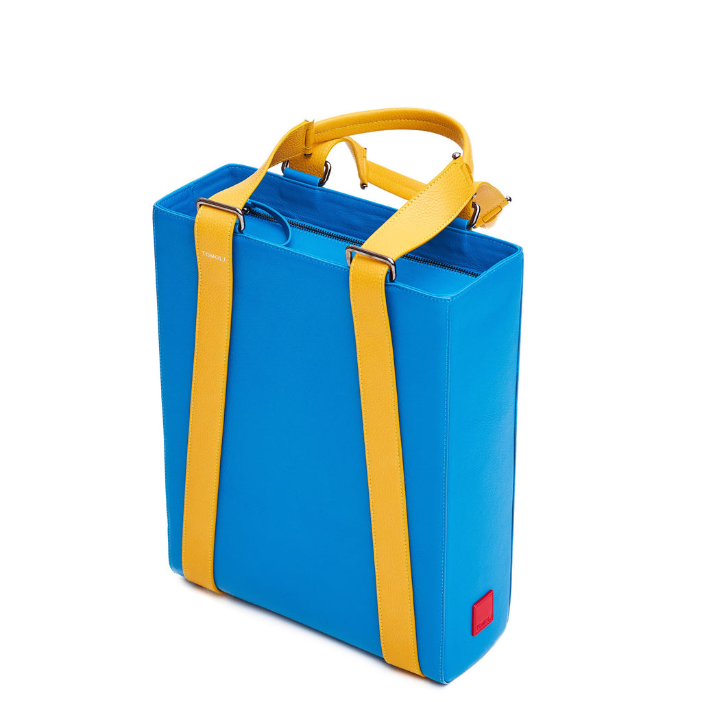 The product photo of a turquoise blue leather tote bag. The tote bag has a structured rectangular briefcase shape and is color-blocked with yellow leather straps. This is the side view of the Tomoli Kora large convertible leather tote in Golden Sky. The bag can fit a laptop and can be styled for work or to elevate casual outfits.