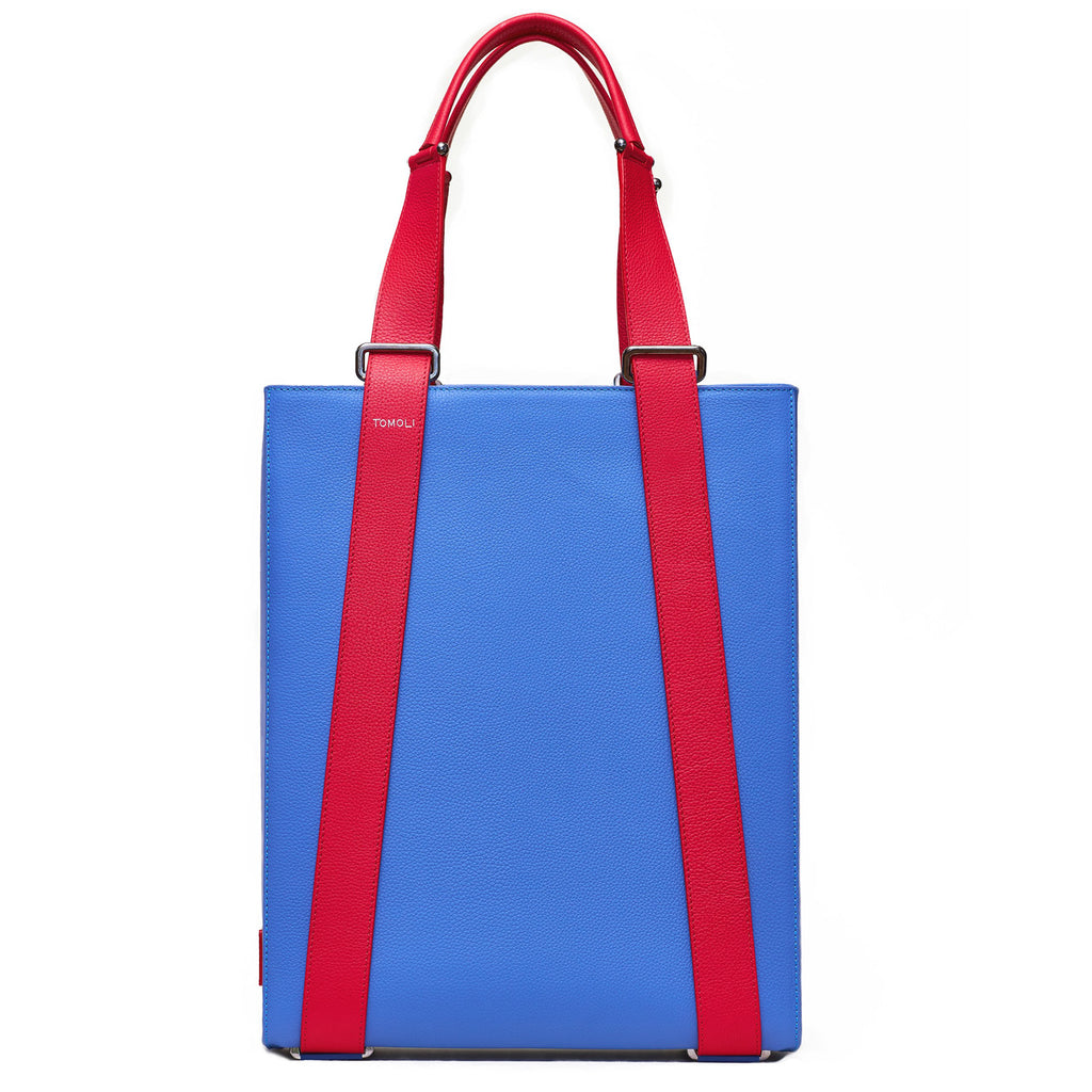 The product photo of a blue purple leather tote bag. The tote bag has a structured rectangular briefcase shape and is color-blocked with red leather straps. This is the front view of the Tomoli Kora large convertible leather tote in Hot Iris. The bag can fit a laptop and can be styled for work or to elevate casual outfits.