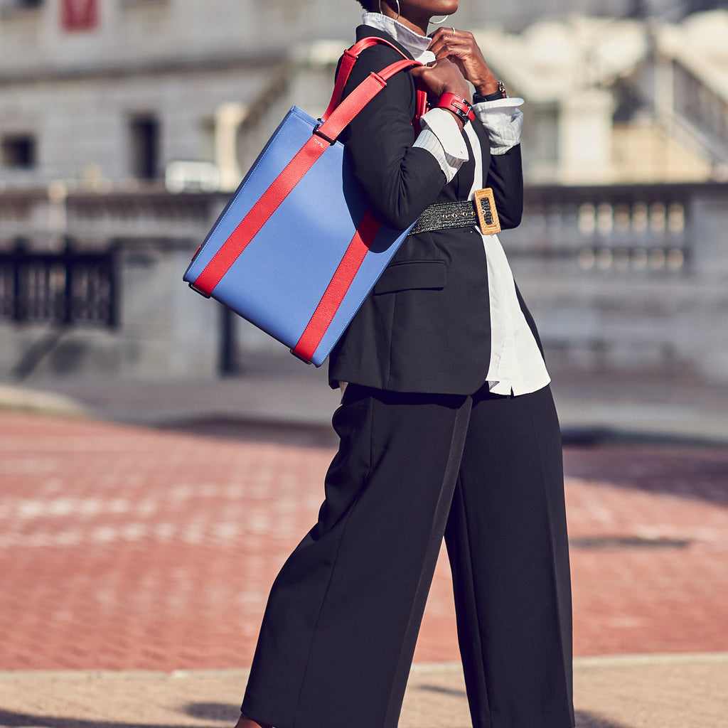 An outfit and style inspiration photo on how to wear a large tote bag. A woman wearing a black and white outfit is holding a large blue purple leather tote bag. The handbag has a structured rectangular shape and is color-blocked with red straps. This is the Tomoli Kora large convertible leather tote in Hot Iris.