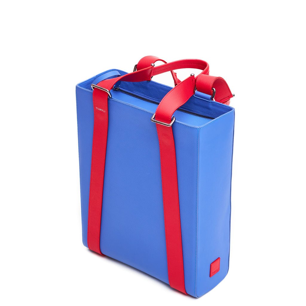 The product photo of a blue purple leather tote bag. The tote bag has a structured rectangular briefcase shape and is color-blocked with red leather straps. This is the side view of the Tomoli Kora large convertible leather tote in Hot Iris. The bag can fit a laptop and can be styled for work or to elevate casual outfits.