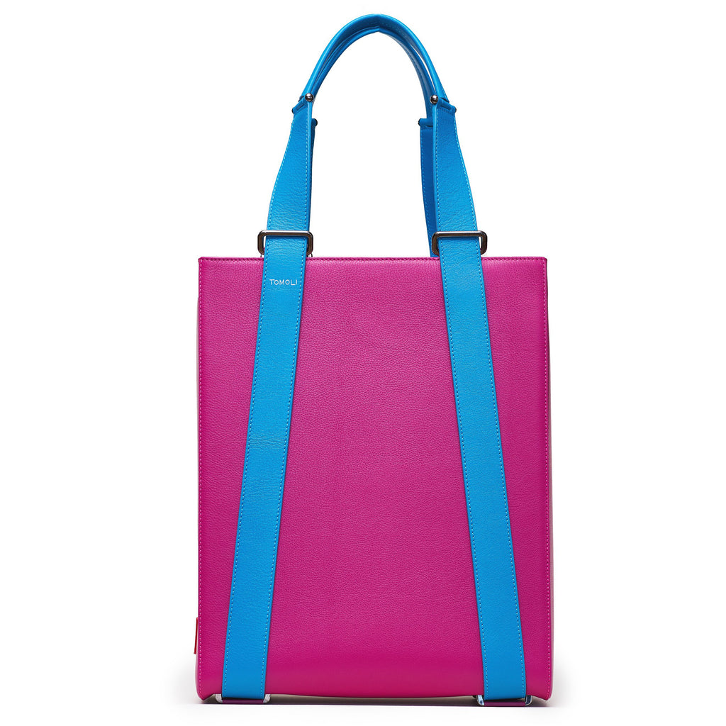 The product photo of a pink leather tote bag. The tote bag has a structured rectangular briefcase shape and is color-blocked with blue leather straps. This is the front view of the Tomoli Kora large convertible leather tote in sky fuchsia. The bag can fit a laptop and can be styled for work or to elevate casual outfits.
