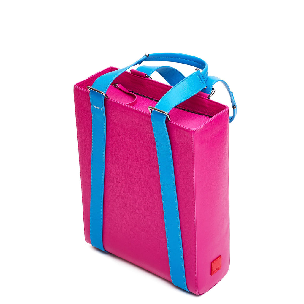 The product photo of a pink leather tote bag. The tote bag has a structured rectangular briefcase shape and is color-blocked with blue leather straps. This is the side view of the Tomoli Kora large convertible leather tote in sky fuchsia. The bag can fit a laptop and can be styled for work or to elevate casual outfits.