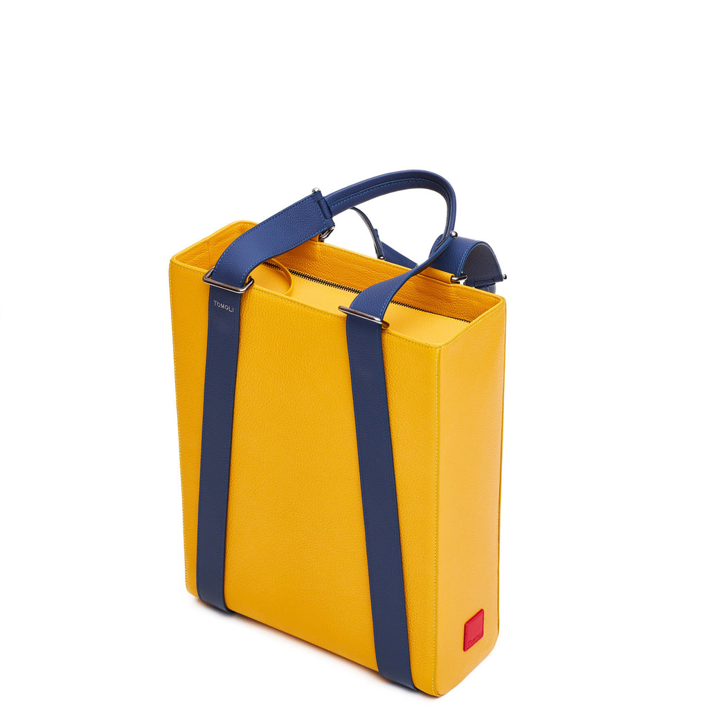 The product photo of a colorful yellow leather tote bag. The tote bag has a structured rectangular briefcase shape and is color-blocked with dark blue leather straps. This is the side view of the Tomoli Kora large convertible leather tote in shady maize. The bag can fit a laptop and can be styled for work or to elevate casual outfits.