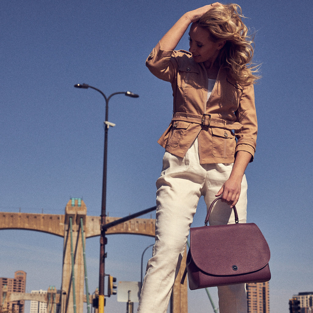 A fashion and style editorial photo showing a model holding a burgundy red leather handbag. The bag has a minimalistic design and an oversized round flap cover. This is the Tomoli Briffani Jut interchangeable satchel in Sangria.