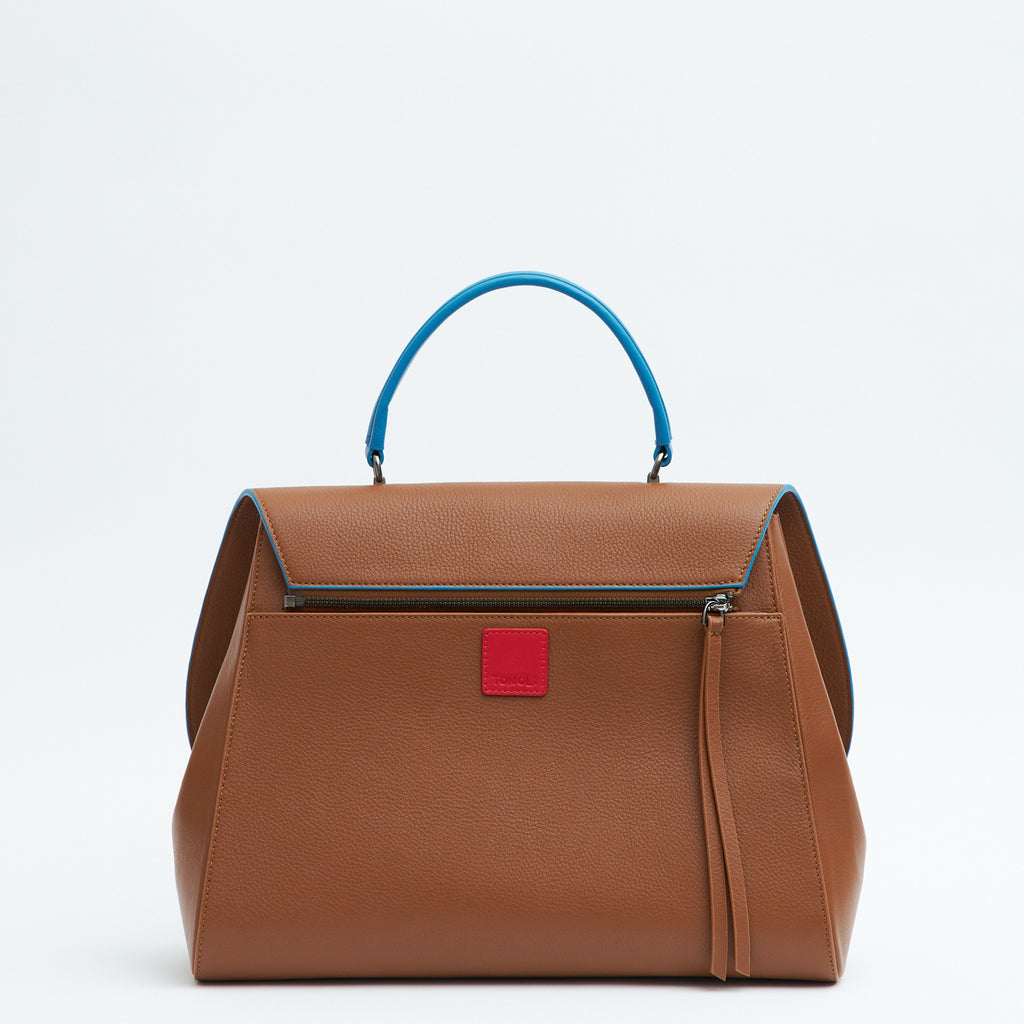 A product photo showing the back of a brown top handle leather handbag. The handbag has a minimalistic design and is color-blocked with a blue top handle. The back of the bag has a zipper attachment with a small red square underneath it. This is the Tomoli Briffani Jut interchangeable satchel in Sky Pecan.