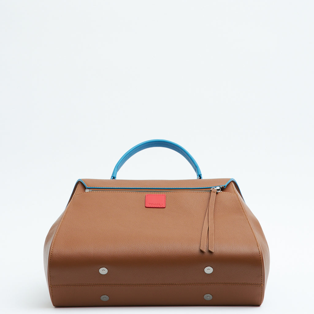 A product photo showing the bottom perspective of a brown top handle leather handbag. The handbag has a minimalistic design and is color-blocked with a blue top handle. The back of the bag has a zipper attachment with a small red square underneath it. This is the Tomoli Briffani Jut interchangeable satchel in Sky Pecan.