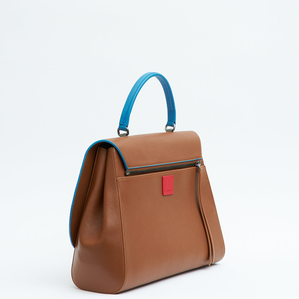A product photo showing the side perspective of a brown top handle leather handbag. The handbag has a minimalistic design and is color-blocked with a blue top handle. The back of the bag has a zipper attachment with a small red square underneath it. This is the Tomoli Briffani Jut interchangeable satchel in Sky Pecan.