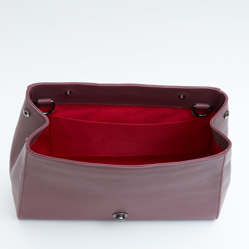 The product photo of a fashion handbag showing the interior of a burgundy red leather bag. The bag has a bright red lining made of alcantara microsuede material. This is the interior view of the Tomoli Briffani Jut handbag in Sangria. This bag fits a laptop and a large iPad Pro tablet.