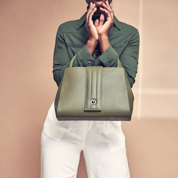 A fashion and style photo showing a model holding an olive green leather tote handbag. The handbag has a classic trapeze shape and a quilted flap closure. This is the Tomoli Briffani Lean interchangeable tote handbag in Olive Green.