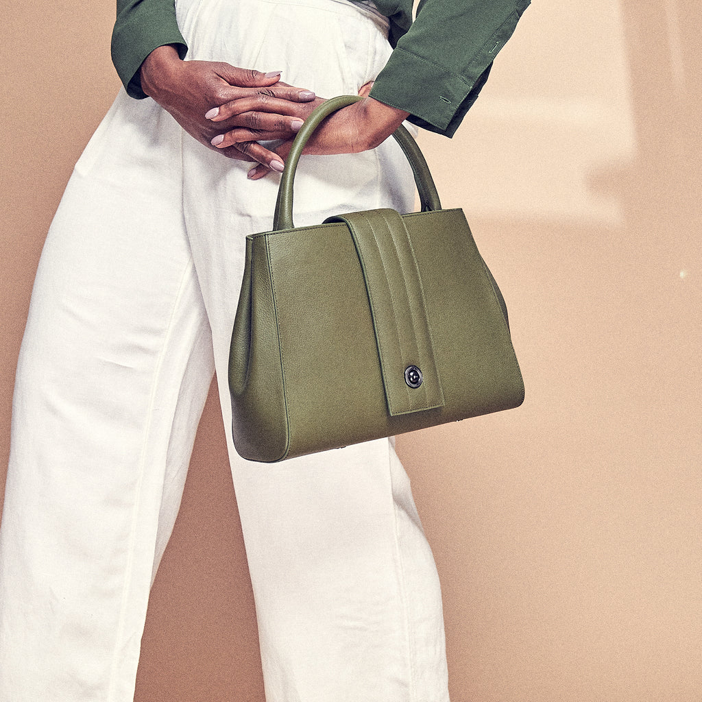 A fashion and style photo showing a model holding an olive green leather tote handbag. The handbag has a classic trapeze shape and a quilted flap closure. This is the Tomoli Briffani Lean interchangeable tote handbag in Olive Green. This bag can be used as an everyday bag, a work tote, or an oversized clutch.