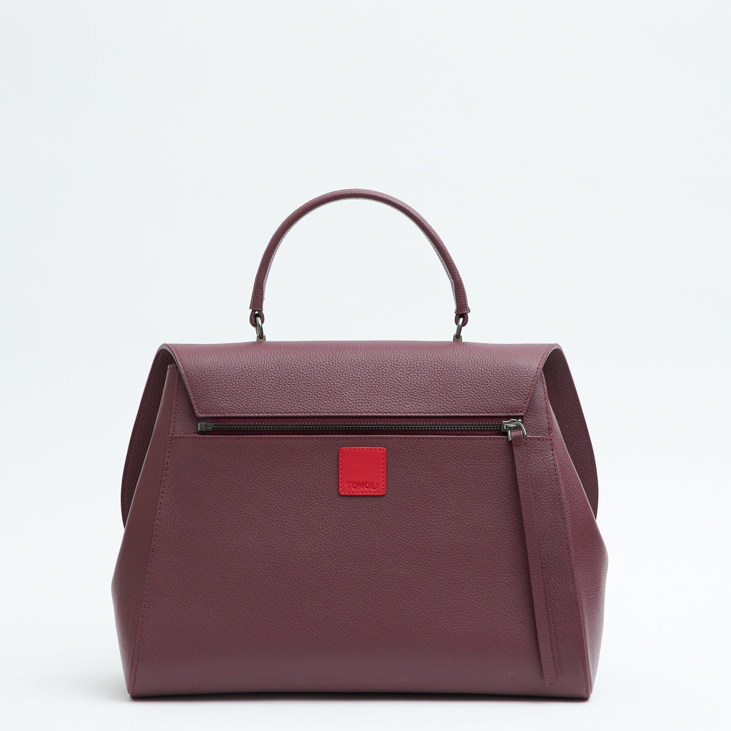 A product photo showing the back of a burgundy red top handle leather handbag. The handbag has a minimalistic design and a zipper attachment. There is a small red square underneath the zipper. This is the Tomoli Briffani Jut interchangeable satchel in Sangria.