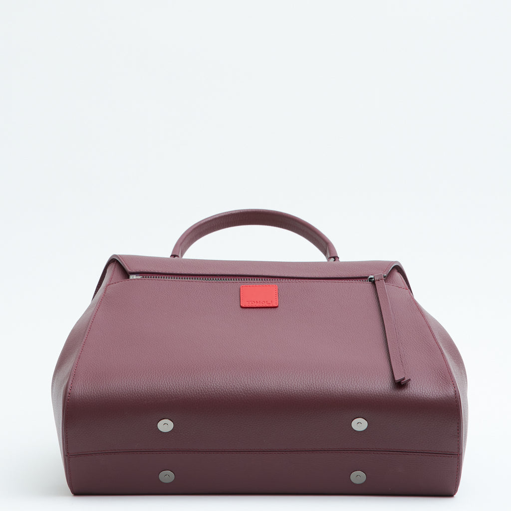 A product photo showing the bottom perspective of a burgundy red top handle leather handbag. The handbag has a minimalistic design and a zipper attachment in the back. There is a small red square underneath the zipper. This is the Tomoli Briffani Jut interchangeable satchel in Sangria.