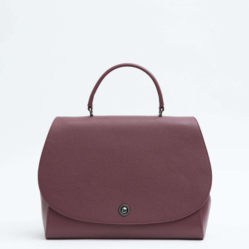The product photo of a burgundy red top handle leather handbag. The handbag has a minimalistic design and an oversized round flap cover. This is the Tomoli Briffani Jut interchangeable satchel  in Sangria.