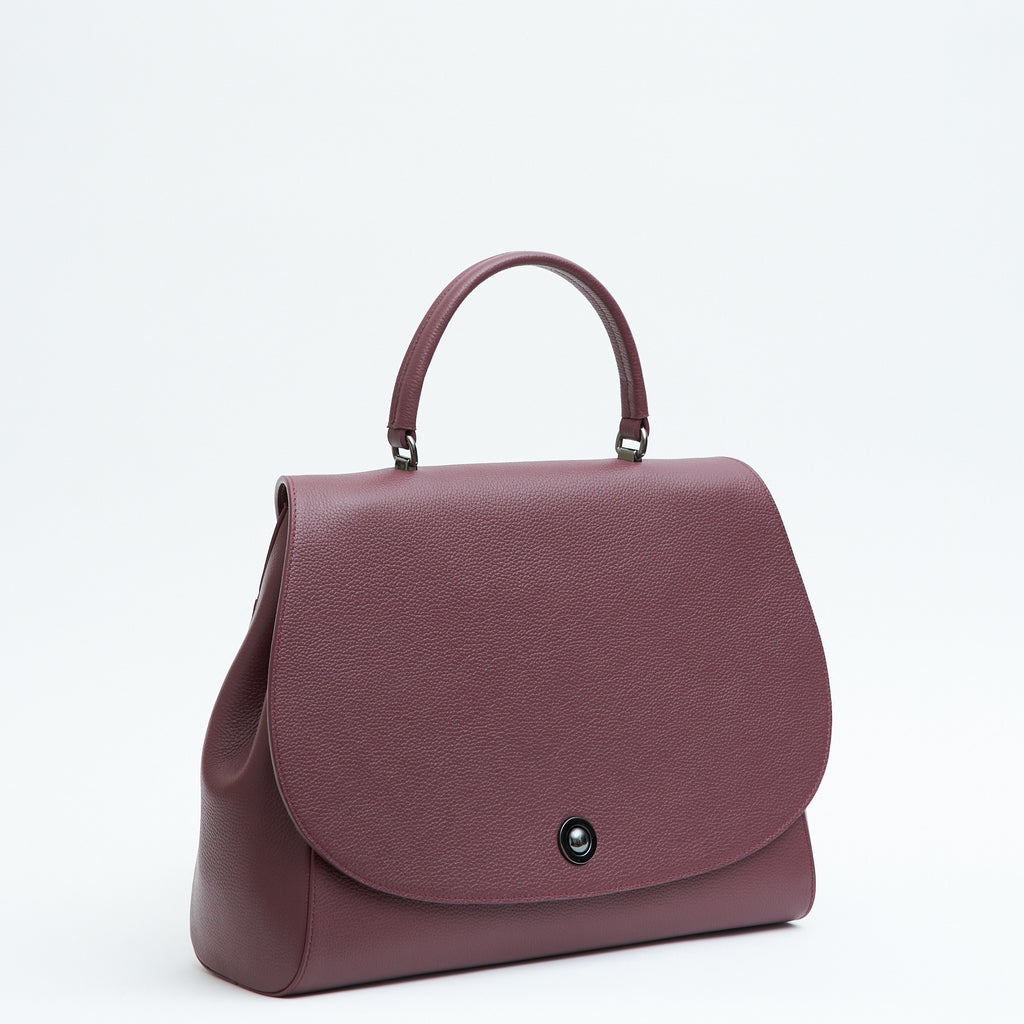 A product photo showing the side of a burgundy red top handle leather handbag. The handbag has a minimalistic design and an oversized round flap cover. This is the Tomoli Briffani Jut interchangeable satchel in Sangria.