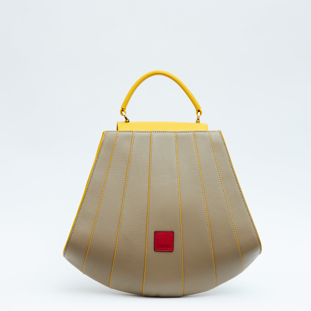 A product photo showing the back of a colorful beige leather handbag. The bag has a tapered trapeze shape that looks like a seashell. The bottom of the bag is rounded and there are yellow lines that create a color-blocked look. This is the Tomoli Gisel tapered top handle handbag in Sun Stone.