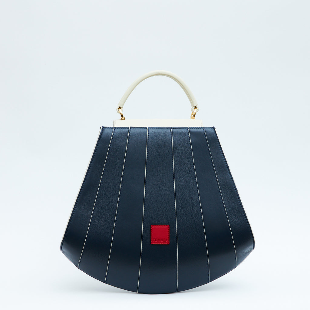 A product photo showing the back of a colorful blue and off-white leather handbag. The bag has a tapered trapeze shape that looks like a seashell. The bottom of the bag is rounded and there are off-white lines that create a color-blocked look. This is the Tomoli Gisel tapered top handle handbag in Ivorian Denim.