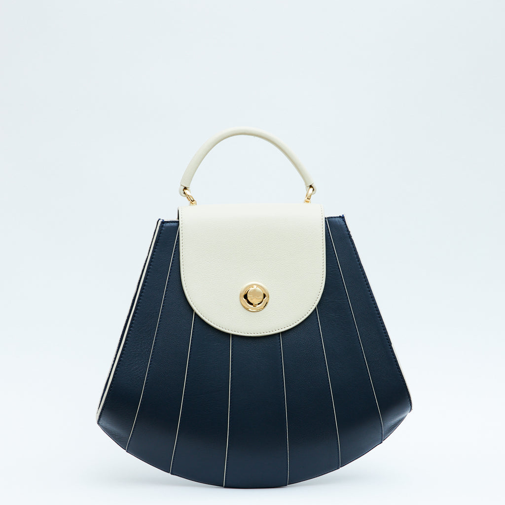 The product photo of a colorful blue and off-white leather handbag. The bag has a tapered trapeze shape that looks like a seashell. The bottom of the bag is rounded and there are off-white radial lines and an off-white flap cover that create a color-blocked look. This is the Tomoli Gisel tapered top handle handbag in Ivorian Denim.