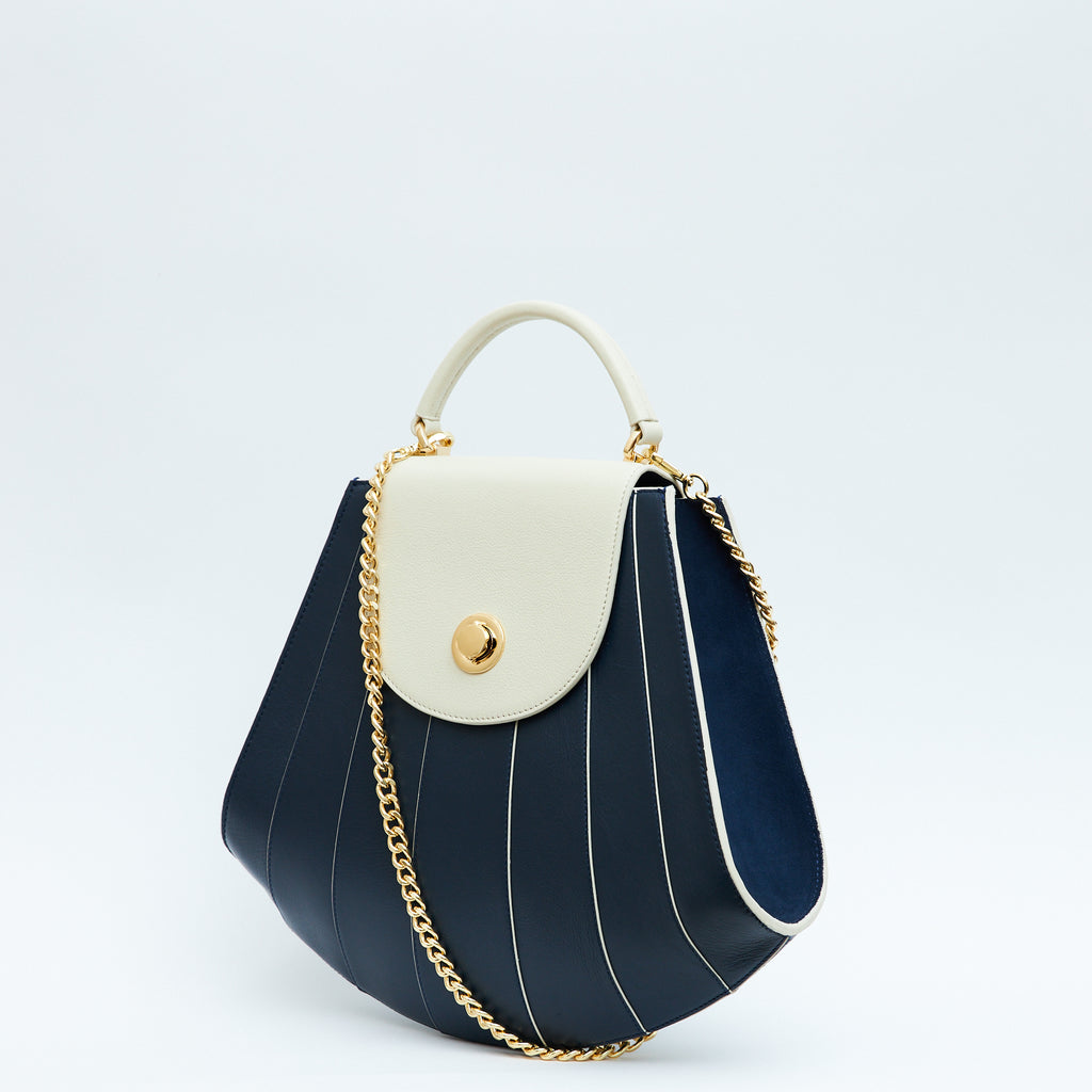A product photo showing the side perspective of a colorful blue and off-white leather handbag. The bag has a tapered trapeze shape that looks like a seashell. The bottom of the bag is rounded and there are off-white radial lines and an off-white flap cover that create a color-blocked look. The bag has a gold chain strap. This is the Tomoli Gisel tapered top handle handbag in Ivorian Denim.