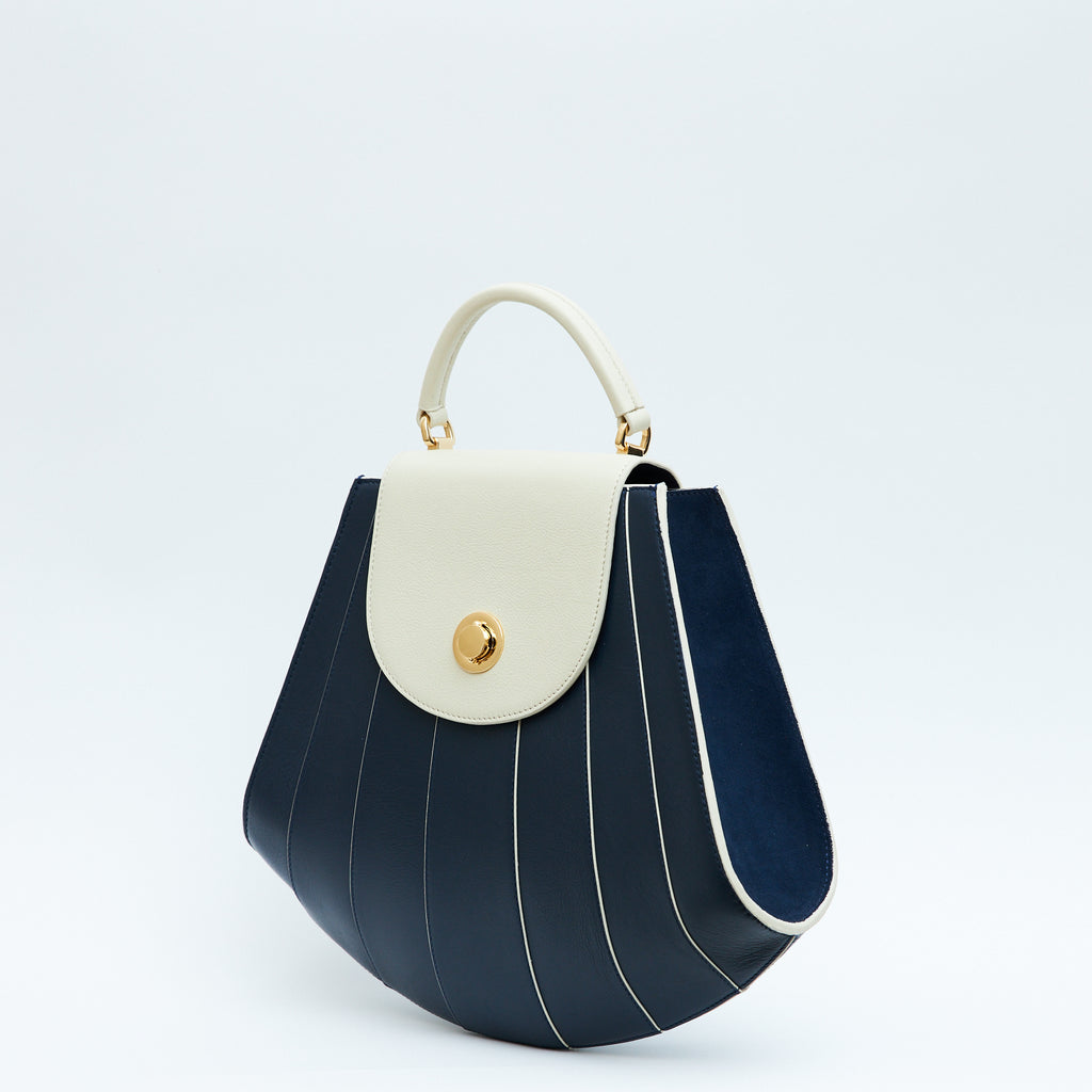 The product photo showing the side perspective of a colorful blue and off-white leather handbag. The bag has a tapered trapeze shape that looks like a seashell. The bottom of the bag is rounded and there are off-white radial lines and an off-white flap cover that create a color-blocked look. This is the Tomoli Gisel tapered top handle handbag in Ivorian Denim.