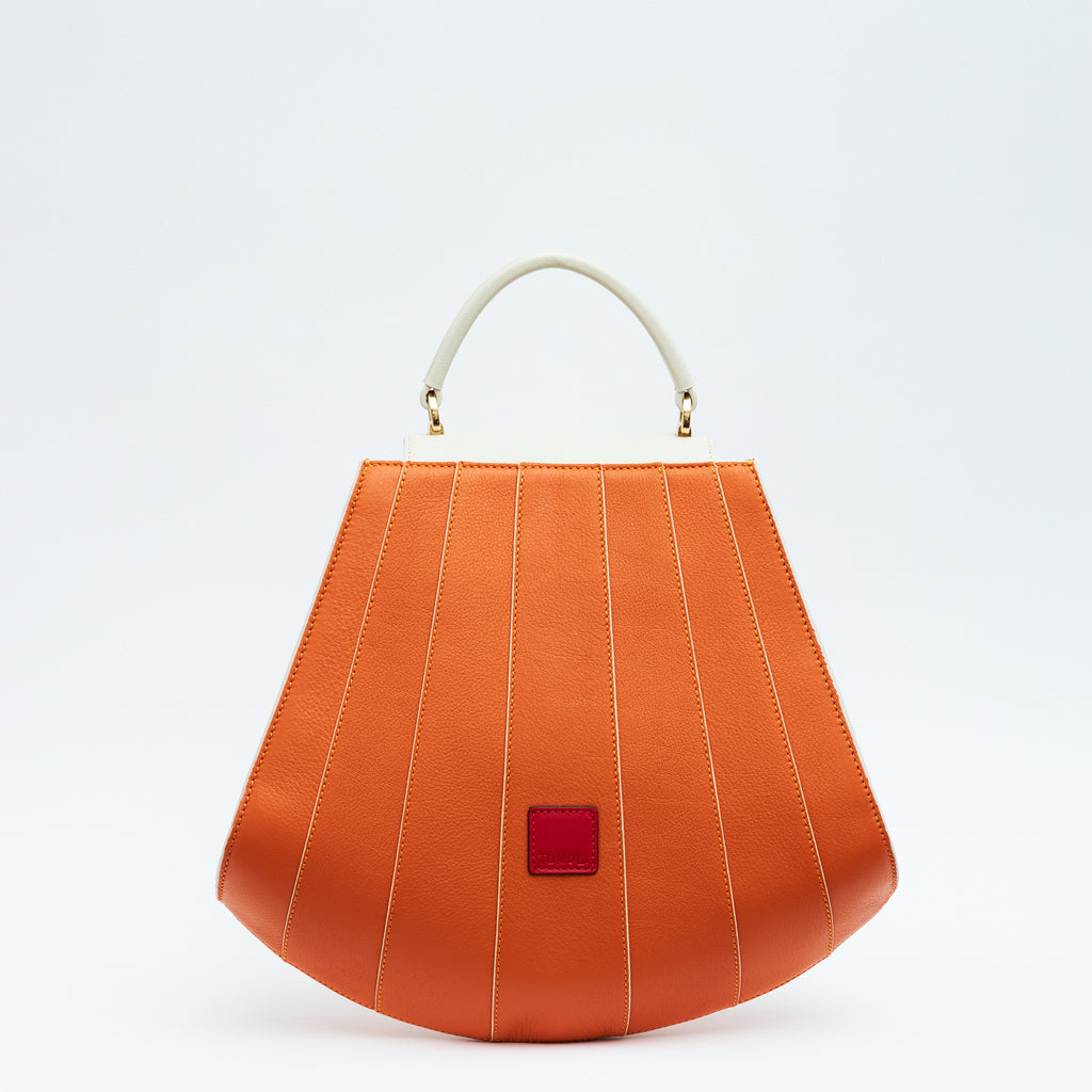 A product photo showing the back of a colorful orange and off-white leather handbag. The bag has a tapered trapeze shape that looks like a seashell. The bottom of the bag is rounded and there are off-white lines that create a color-blocked look. This is the Tomoli Gisel tapered top handle handbag in Ivorian Blaze.