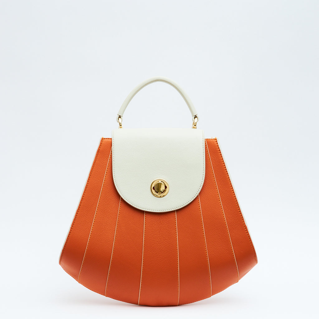 The product photo of a colorful orange and off-white leather handbag. The bag has a tapered trapeze shape that looks like a seashell. The bottom of the bag is rounded and there are off-white contrasting lines and an off-white flap cover that create a color-blocked look. This is the Tomoli Gisel tapered top handle handbag in Ivorian Blaze.