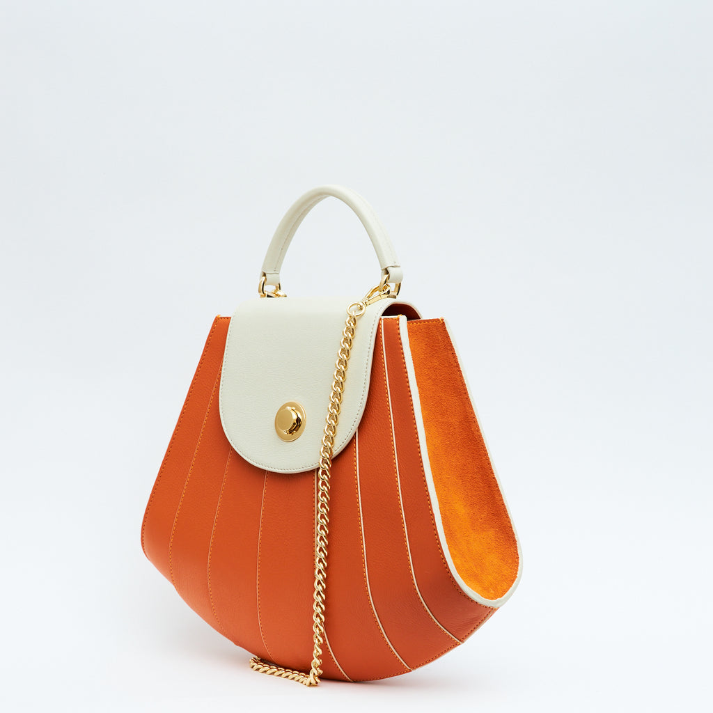 A product photo showing the side perspective of a colorful orange and off-white leather handbag. The bag has a tapered trapeze shape that looks like a seashell. The bottom of the bag is rounded and there are off-white radial lines and an off-white flap cover that create a color-blocked look. The bag has a gold chain strap. This is the Tomoli Gisel tapered top handle handbag in Ivorian Blaze.