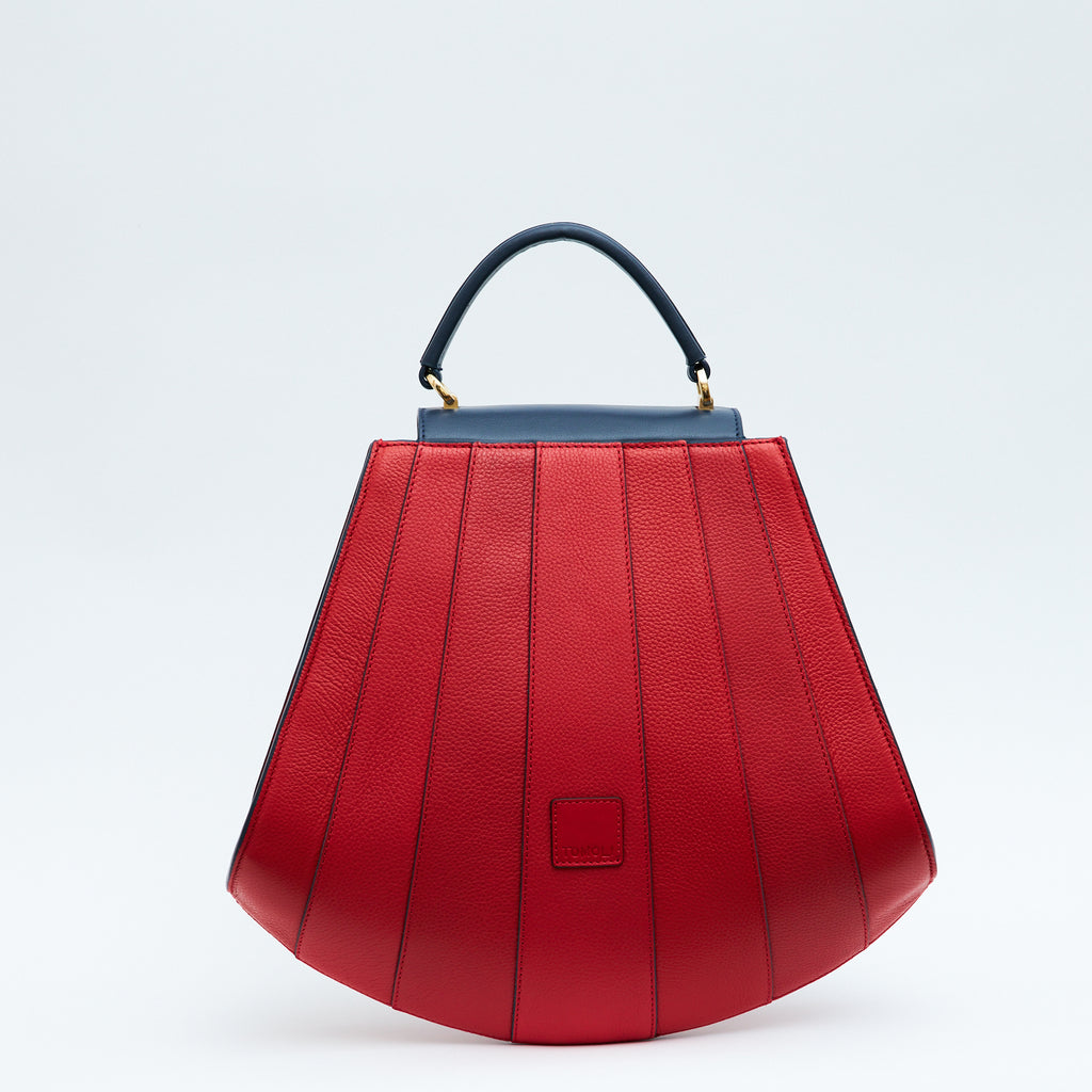 A product photo showing the back of a colorful red leather handbag. The bag has a tapered trapeze shape that looks like a seashell. The bottom of the bag is rounded and there are dark blue lines that create a color-blocked look. This is the Tomoli Gisel tapered top handle handbag in Shady Red.