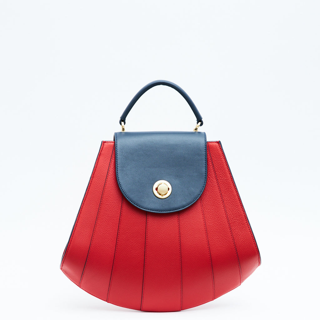 The product photo of a colorful red leather handbag. The bag has a tapered trapeze shape that looks like a seashell. The bottom of the bag is rounded and there are dark blue radial lines and a dark blue flap cover that create a color-blocked look. This is the Tomoli Gisel tapered top handle handbag in Shady Red.