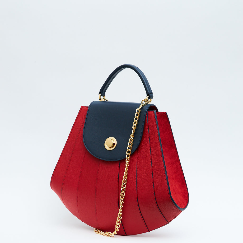 A product photo showing the side perspective of a colorful red leather handbag. The bag has a tapered trapeze shape that looks like a seashell. The bottom of the bag is rounded and there are dark blue radial lines and a dark blue flap cover that create a color-blocked look. The bag has a gold chain strap. This is the Tomoli Gisel tapered top handle handbag in Shady Red.