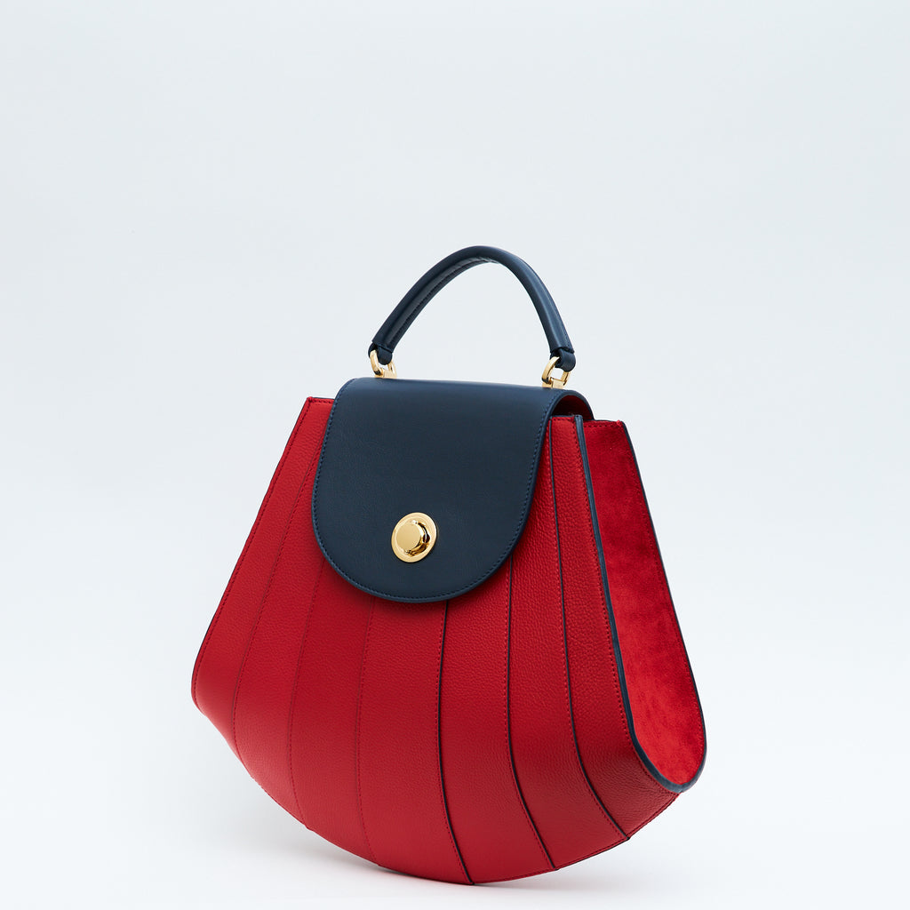 The product photo showing the side perspective of a colorful red leather handbag. The bag has a tapered trapeze shape that looks like a seashell. The bottom of the bag is rounded and there are dark blue radial lines and a dark blue flap cover that create a color-blocked look. This is the Tomoli Gisel tapered top handle handbag in Shady Red.