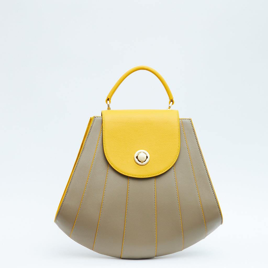 The product photo of a colorful beige leather handbag. The bag has a tapered trapeze shape that looks like a seashell. The bottom of the bag is rounded and there are yellow radial lines and a yellow flap cover that create a color-blocked look. This is the Tomoli Gisel tapered top handle handbag in Sun Stone.