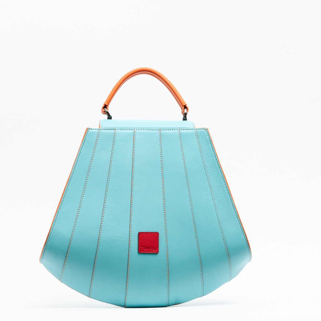 A product photo showing the back of a colorful orange and marine blue leather handbag. The bag has a tapered trapeze shape that looks like a seashell. The bottom of the bag is rounded and there are orange radial lines and an orange handle that create a color-blocked look. This is the Tomoli Gisel tapered top handle handbag in Blazing Marine.