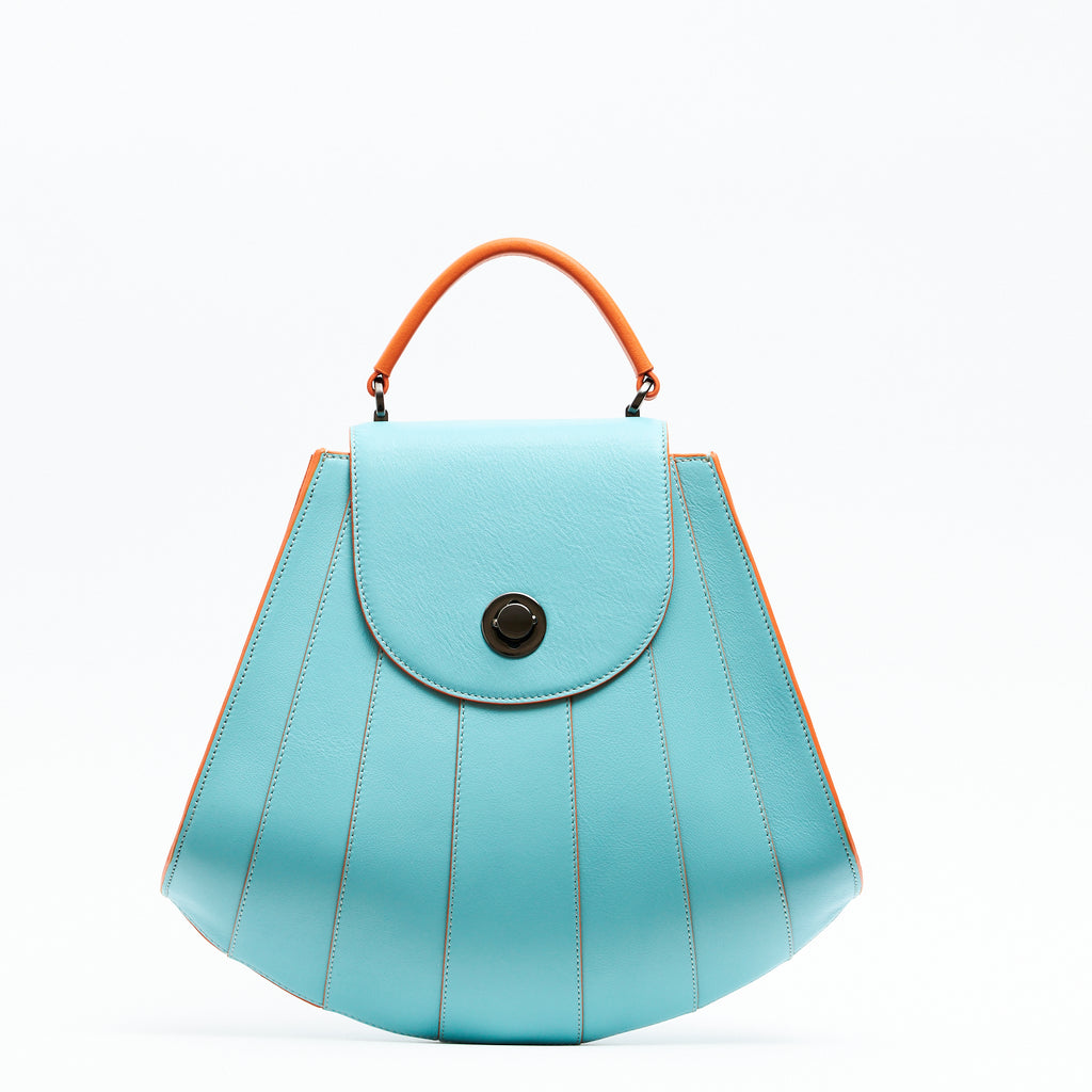 The product photo of a colorful orange and marine blue leather handbag. The bag has a tapered trapeze shape that looks like a seashell. The bottom of the bag is rounded and there are orange radial lines and an orange handle that create a color-blocked look. This is the Tomoli Gisel tapered top handle handbag in Blazing Marine.