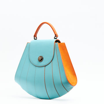 The product photo showing the side perspective of a colorful orange and marine blue leather handbag. The bag has a tapered trapeze shape that looks like a seashell. The bottom of the bag is rounded and there are orange radial lines that create a color-blocked look. This is the Tomoli Gisel tapered top handle handbag in Blazing Marine.