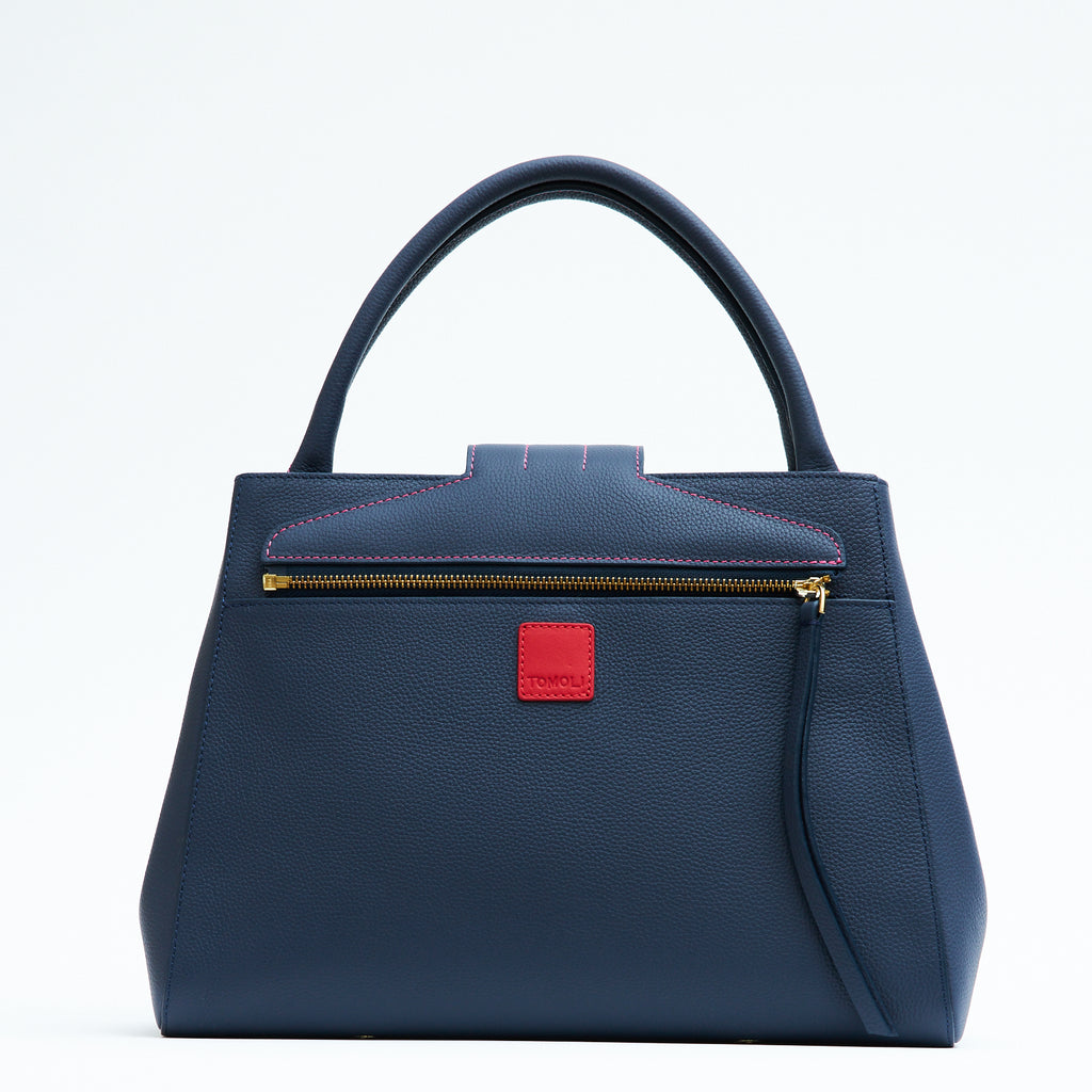 A product photo showing the back view of a colorful dark blue leather tote handbag. The handbag has a classic trapeze shape with dark blue top handles, a back pocket, and a zipper to interchange covers. This is the Tomoli Briffani Lean interchangeable tote handbag in Flirty Denim. This bag can be used as an everyday bag, a work tote, or even an oversized clutch.