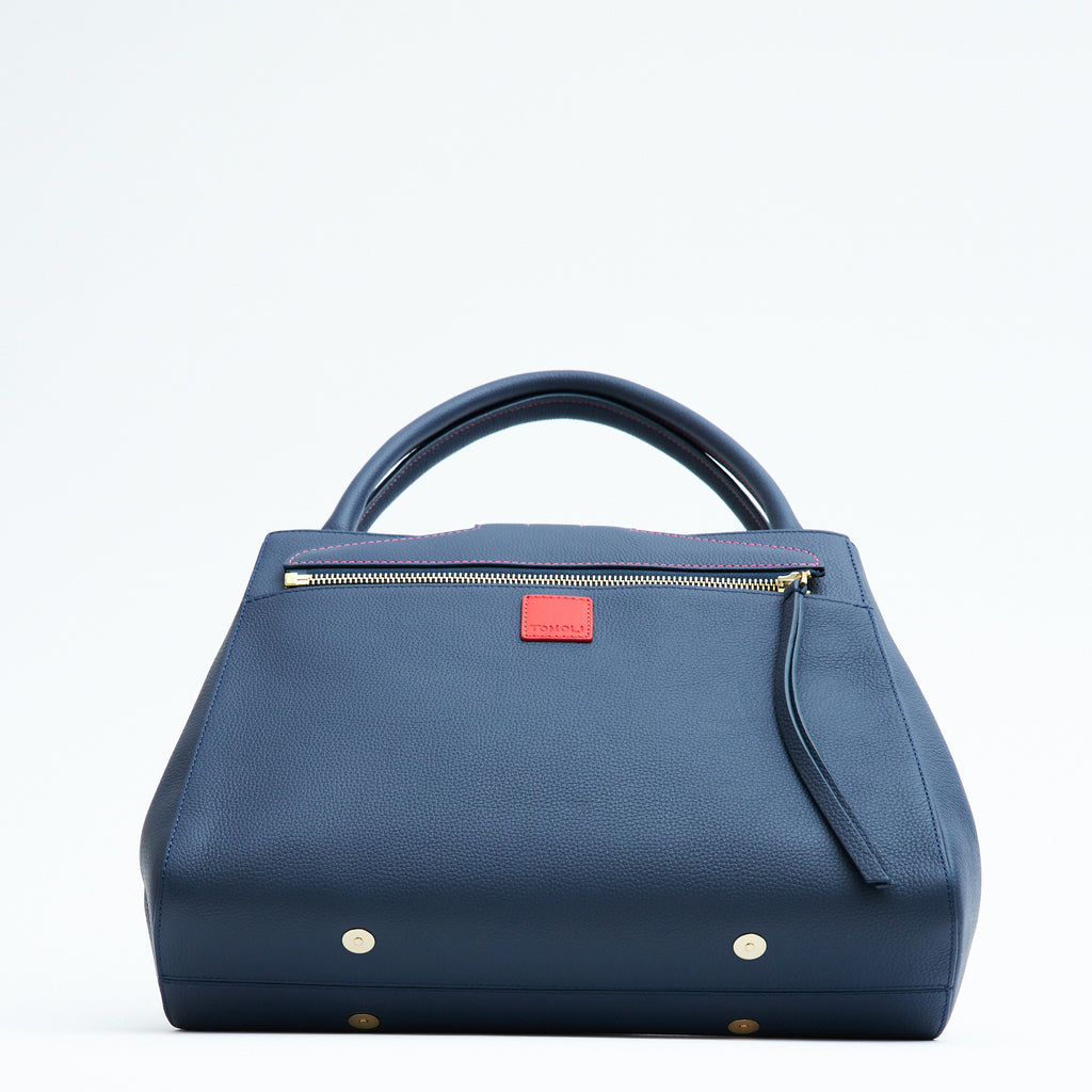 A product photo showing the bottom perspective view of a colorful dark blue leather tote handbag. The handbag has a classic trapeze shape with dark blue top handles, a back pocket, and a zipper to interchange covers. There are four gold rivets at the bottom. This is the Tomoli Briffani Lean interchangeable tote handbag in Flirty Denim. This bag can be used as an everyday bag, a work tote, or even an oversized clutch.
