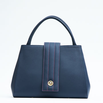 A product photo showing the front view of a colorful dark blue leather tote handbag. The handbag has a classic trapeze shape with dark blue top handles and a straight quilted flap with contrasting pink stitching. This is the Tomoli Briffani Lean interchangeable tote handbag in Flirty Denim. This fashion bag can be used as an everyday bag, a work tote, or even an oversized clutch.