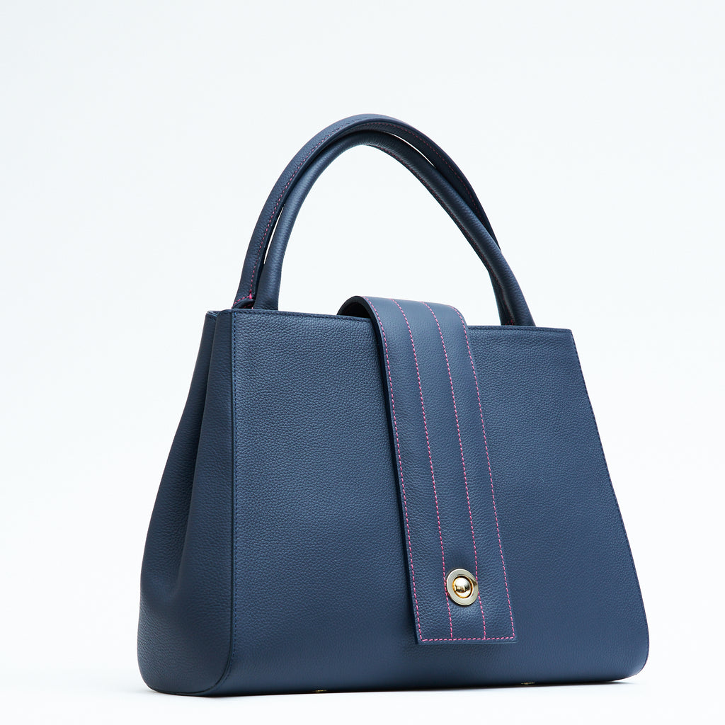 A product photo showing the side view of a colorful dark blue leather tote handbag. The handbag has a classic trapeze shape with dark blue top handles and a straight quilted flap with contrasting pink stitching. This is the Tomoli Briffani Lean interchangeable tote handbag in Flirty Denim. This bag can be used as an everyday bag, a work tote, or even an oversized clutch.