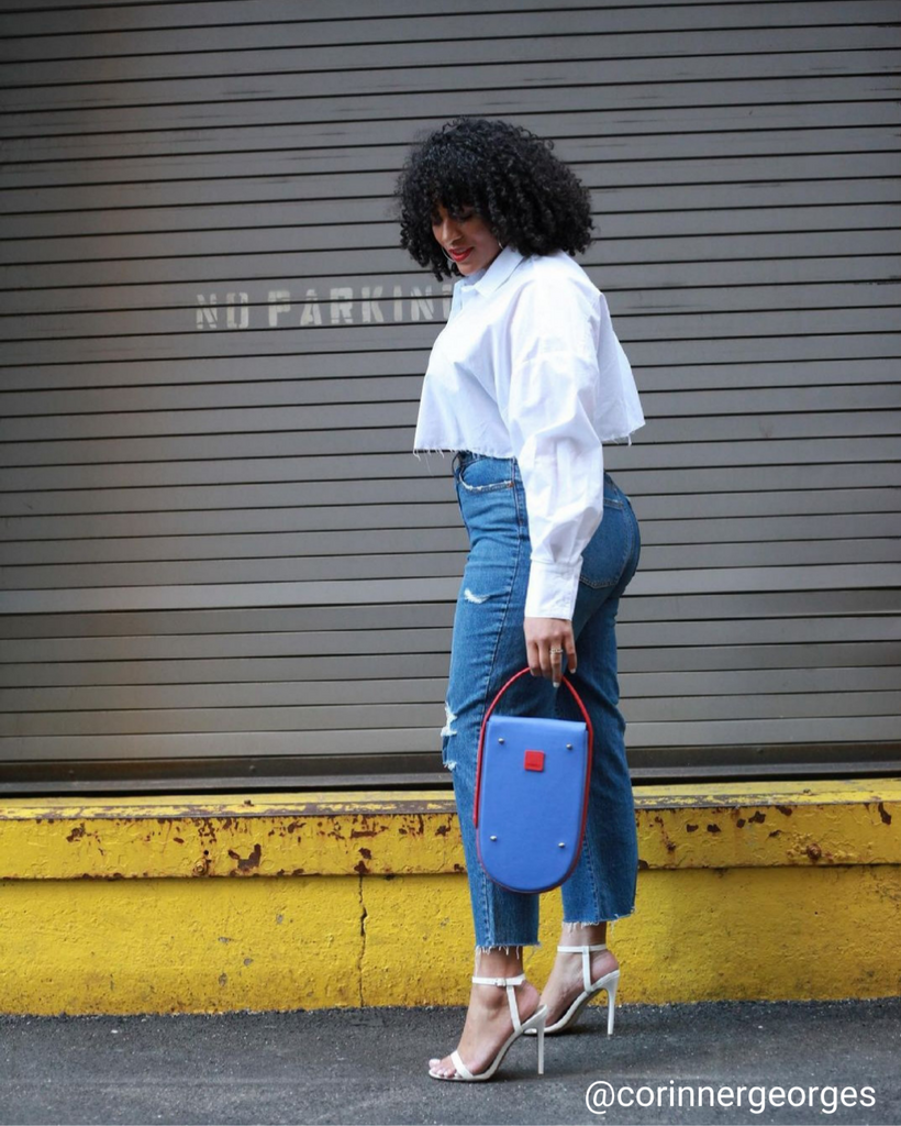 A fashion streetstyle photo showing influencer @corinnergeorges wearing blue jeans and a white shirt and holding a colorful blue purple leather handbag. The handbag has a small, elongated rounded shape. The overall look is color-blocked with white, purple and blue.
