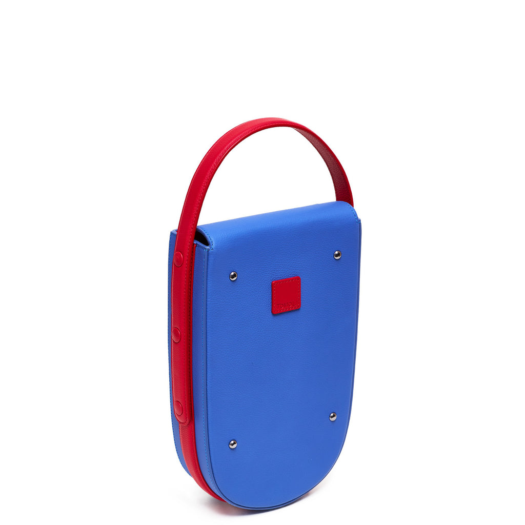 A colorful red and blue mini leather tote handbag. The handbag has an elongated rounded shape and a red adjustable top handle. The hardware is gunmetal. This is the back view of the Tomoli Fitini bag in Hot Iris.