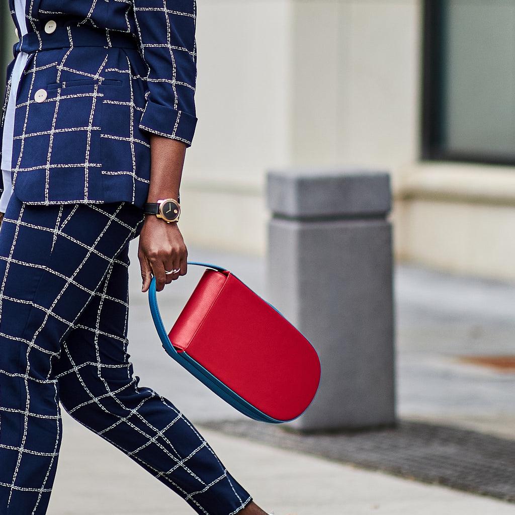 A fashion and style editorial photo showing a woman in a blue pant suit holding a colorful red leather handbag. The handbag has a small, elongated rounded shape.  The overall look is color-blocked with red and blue.