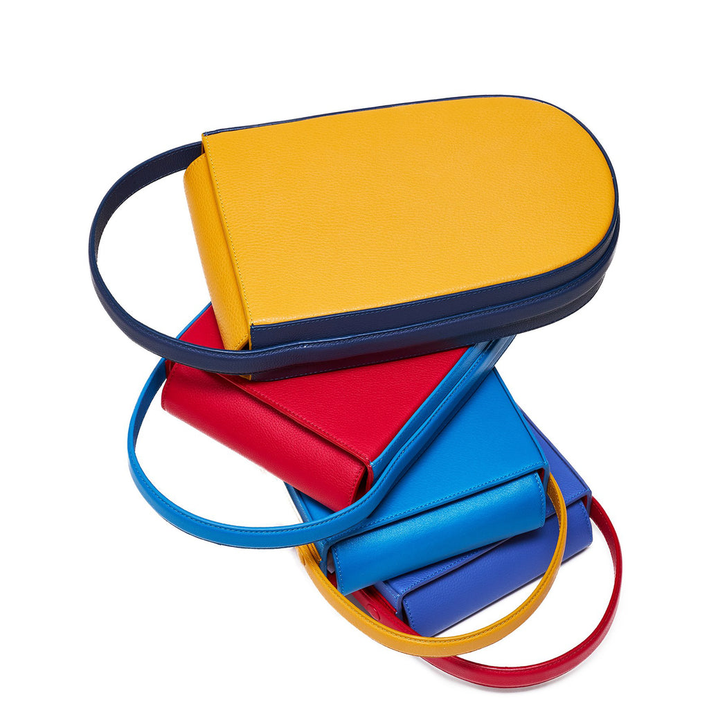 Four colorful fashion tote handbags stacked on top on each other. The handbags all have the same minimalist and elongated rounded shape. There is a yellow tote bag, a red tote bag, an aqua blue tote bag, and a blue purple tote bag. The overall image creates a bold color-blocking effect. This is a group photo of the Tomoli Fitini handbags.