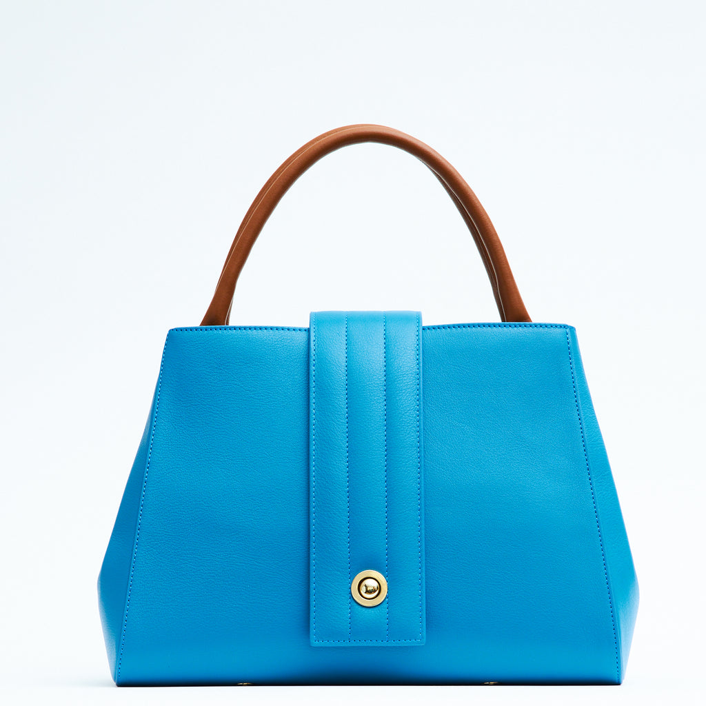 A product photo showing the front view of a colorful turquoise blue leather handbag. The handbag has a classic trapeze shape with brown top handles and a straight quilted flap in the middle. This is the Tomoli Briffani Lean interchangeable tote handbag in Brunette Sky. This bag can be used as an everyday bag, a work tote, or even an oversized clutch.