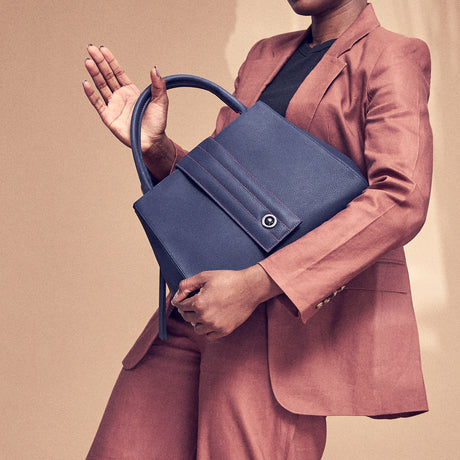 The product photo of a navy blue leather handbag. The handbag has a classic trapeze shape with a quilted flap closure and top handles. This is a front view of the Tomoli Briffani Lean interchangeable tote handbag in Flirty Denim. This bag can be used as an everyday bag, a work tote, or even an oversized clutch.