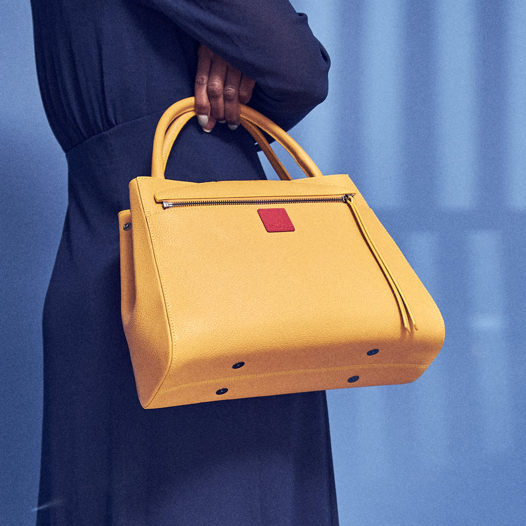 The product photo of a yellow leather handbag. The handbag has a classic trapeze shape with a back pocket, a zipper to interchange flaps, and top handles. This is the back view of the Tomoli Briffani Lean interchangeable tote handbag in Maize. This bag can be used as an everyday bag, a work tote, or even an oversized clutch.