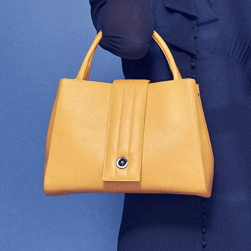 A fashion and style photo showing a model holding a yellow leather handbag. The handbag has a classic trapeze shape with a straight quilted flap in the middle and top handles. This is the front view of the Tomoli Briffani Lean interchangeable tote handbag in Maize. This bag can be used as an everyday bag, a work tote, or even an oversized clutch.