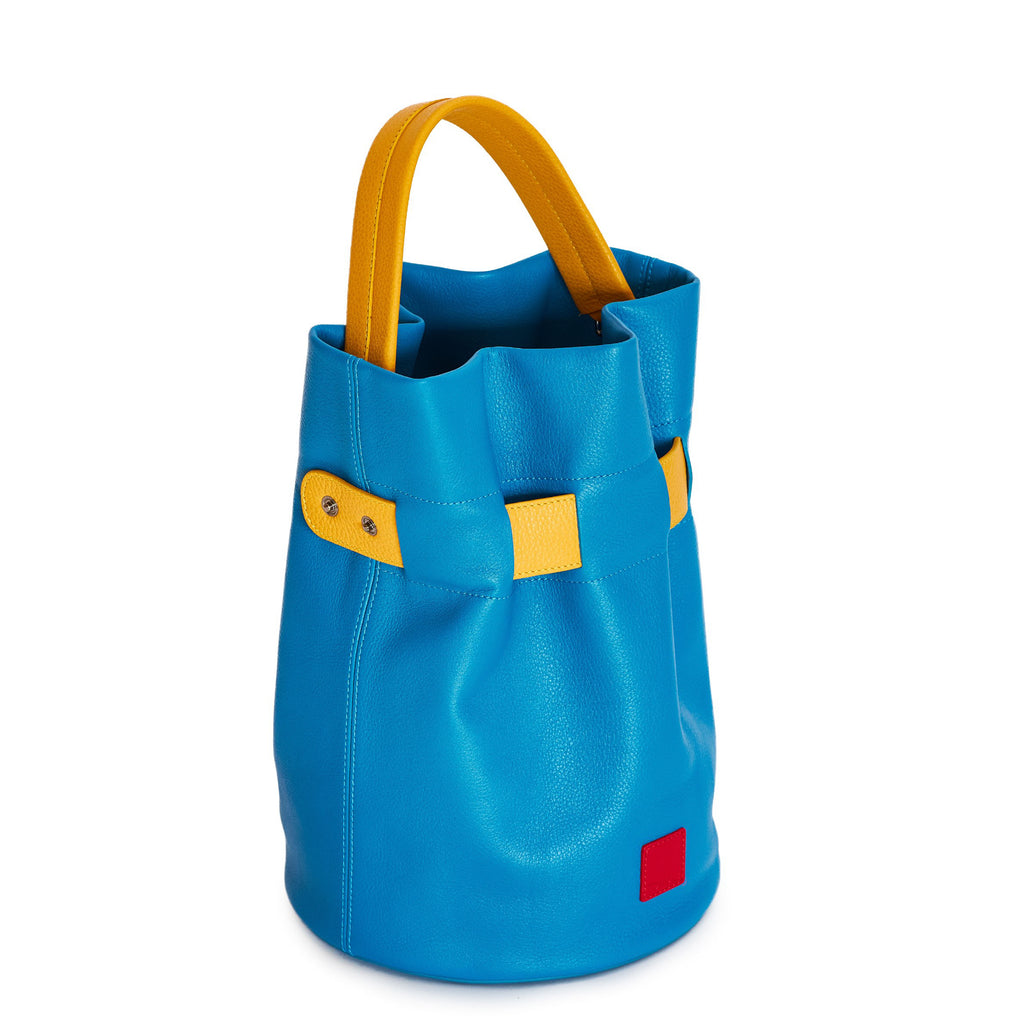 The product photo of a colorful fashion handbag showing a blue leather bucket bag with a yellow belt cinch and a yellow top handle. The belt is woven into the bag and has 2 gunmetal snap buttons. This is the side view of the Tomoli La Bourse leather bucket bag in Golden Sky.