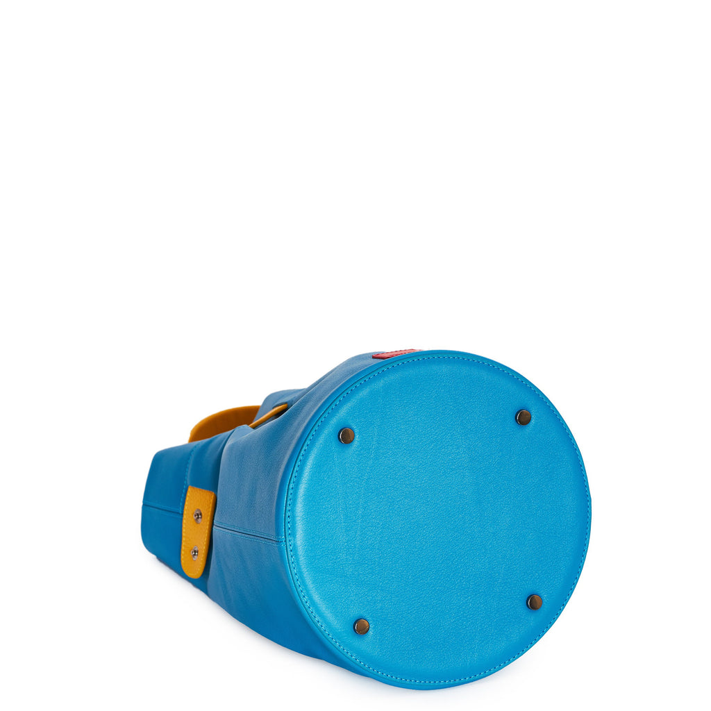 The product photo of a colorful fashion handbag showing a blue leather bucket bag with a yellow belt cinch and a yellow top handle. The bottom of the bag is round and has 4 gunmetal rivets. This is the bottom view of the Tomoli La Bourse leather bucket bag in Golden Sky.