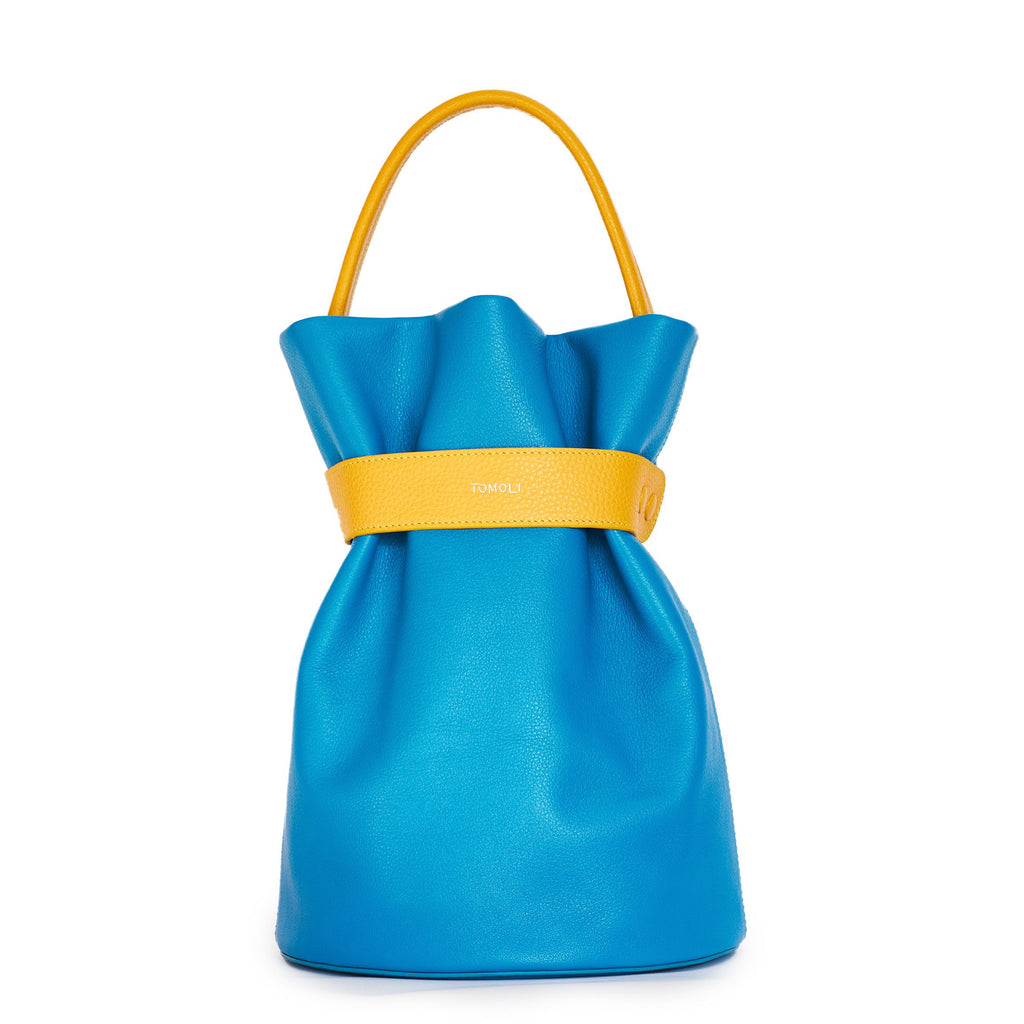 The product photo of a colorful fashion handbag showing a blue leather bucket bag with a yellow belt cinch and a yellow top handle. This is the front view of the Tomoli La Bourse leather bucket bag in Golden Sky.