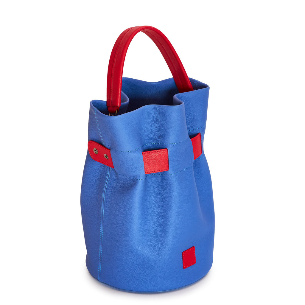 The product photo of a colorful fashion handbag showing a blue leather bucket bag with a red belt cinch and a red top handle. The belt is woven into the bag and has 2 gunmetal snap buttons. This is the side view of the Tomoli La Bourse leather bucket bag in Hot Iris.
